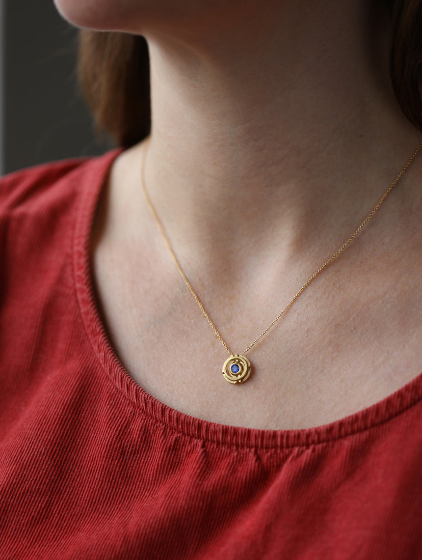 Geometrically patterned pendant featuring a central blue sapphire. Textured 18ct yellow gold. On neck.