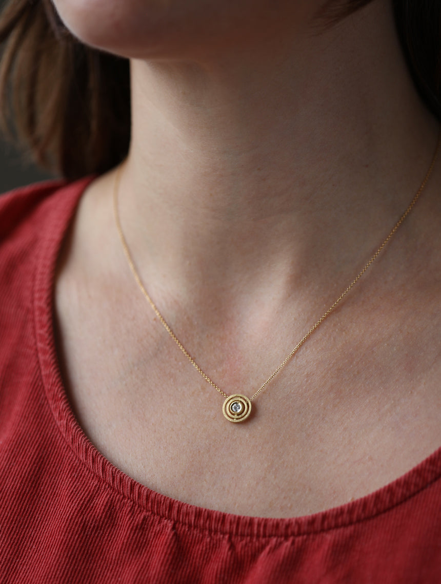 Geometric pendant with concentric circle detail, with central diamond. 18ct yellow gold, textured gold. Worn on neck.