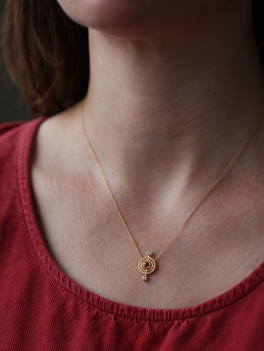 Geometric pendant with circular detail, one orange sapphire and two white diamonds. 18ct yellow gold, textured finish. Worn on neck.